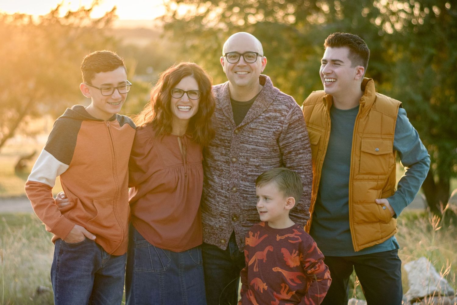 Family of five smiling outdoors at golden hour, parents with three sons posing in a field with warm sunset light.