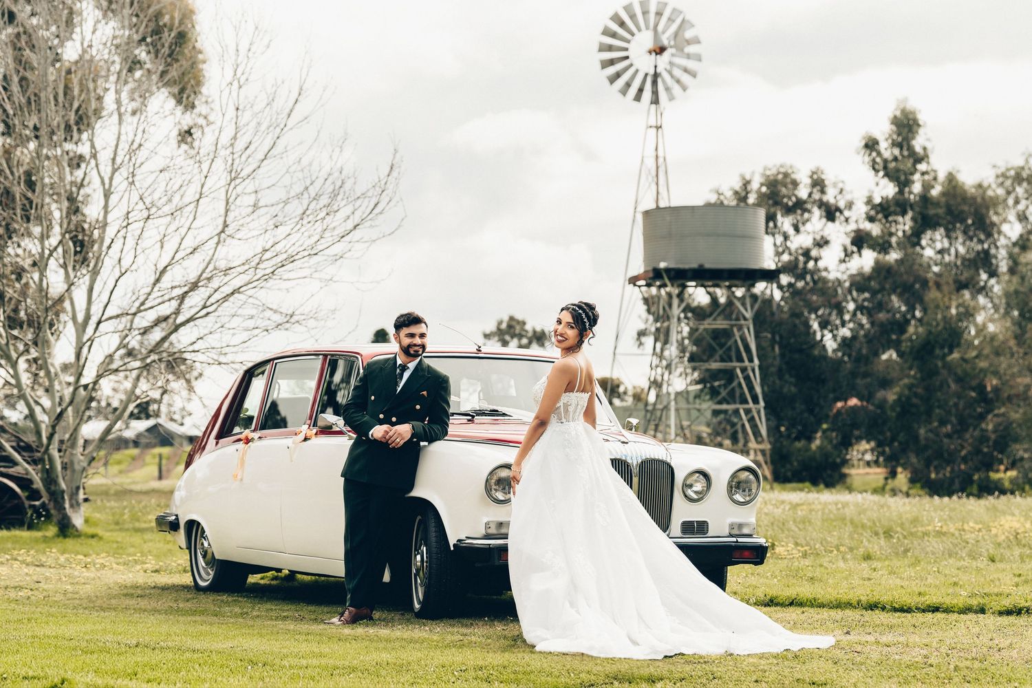Bride in a flowing white gown and groom in a dark suit pose beside a vintage white wedding car in a country field with a windmill.