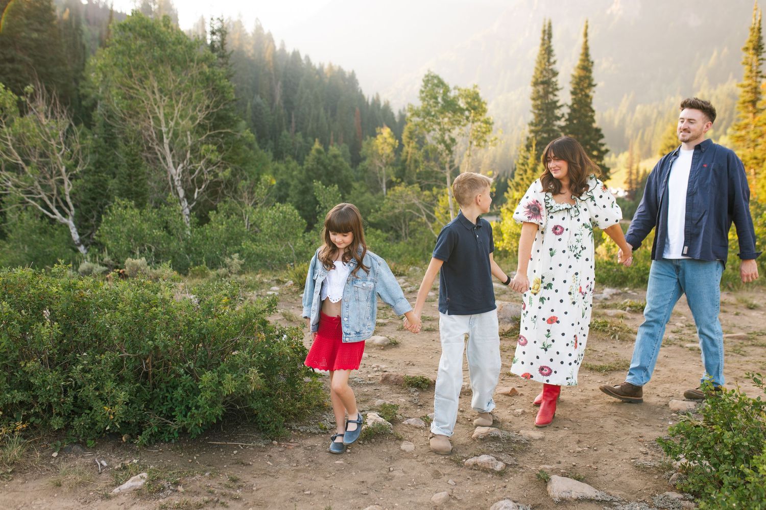 Family of four holding hands walking on a mountain trail surrounded by lush green trees and fall foliage.