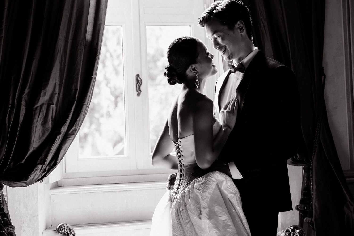 Black and white wedding photo of bride and groom sharing romantic moment by draped window in elegant venue.