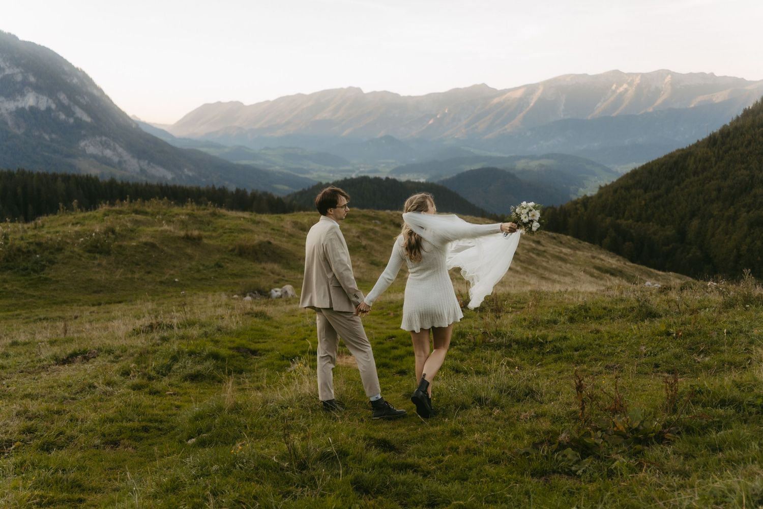 A couple in wedding attire holds hands walking through a mountain meadow, the bride's veil flowing in the wind.