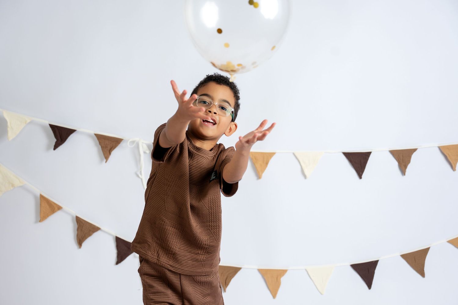 A young boy in glasses and brown outfit joyfully tosses a balloon at a festive party with bunting decorations.