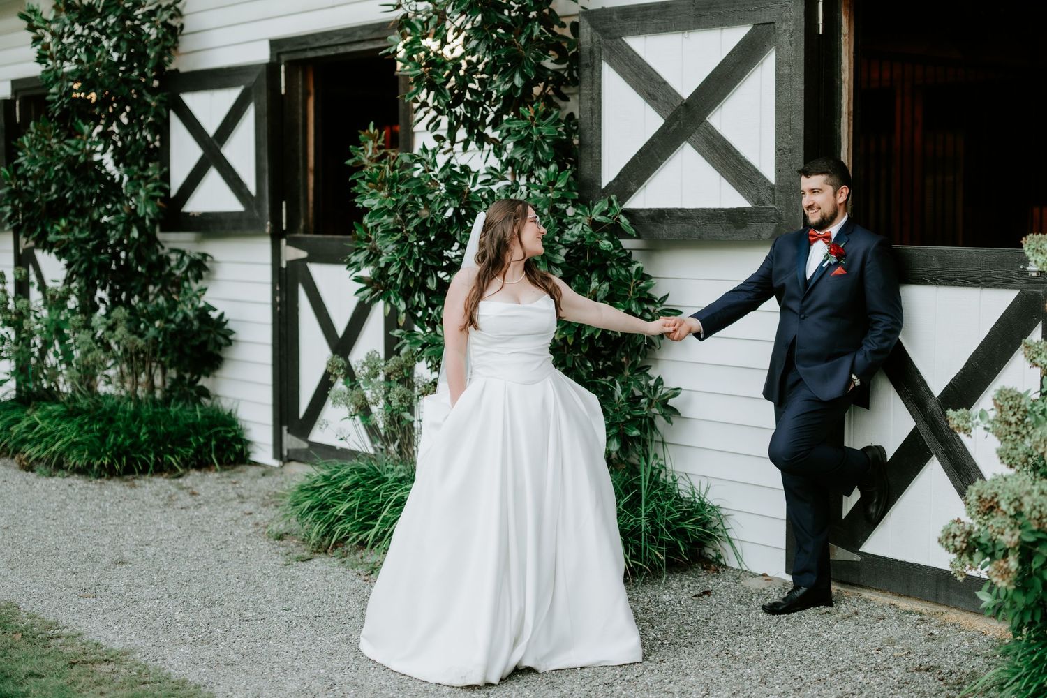Bride and groom hold hands outside a white barn venue, bride in flowing gown, groom in navy suit with red boutonniere.