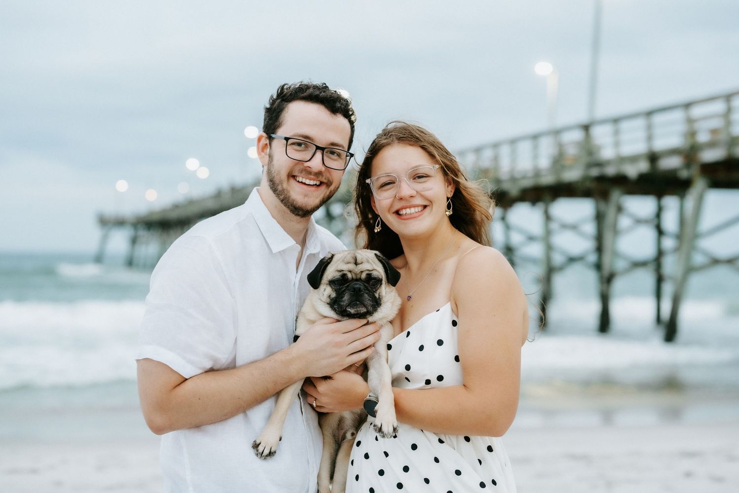 Couple smiling at beach holding a pug dog with ocean pier in background on cloudy day.