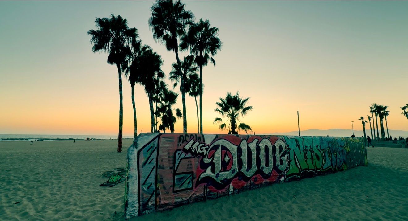 Colorful graffiti mural on Venice Beach at sunset with palm trees silhouetted against golden sky.