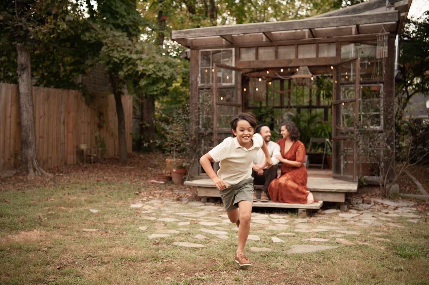 Child running in backyard while mother sits under wooden pergola with baby in lush garden setting.