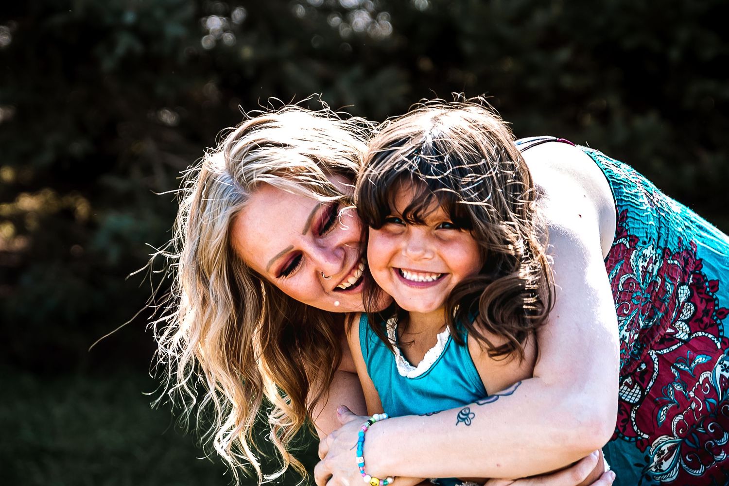 A woman with blonde hair joyfully hugs a laughing young girl outdoors in a lush green garden setting.