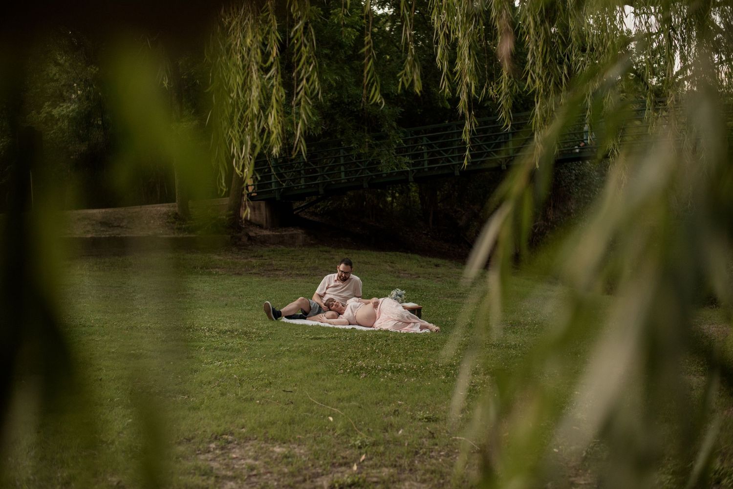 Couple relaxing on a blanket in a lush green park under weeping willow trees.