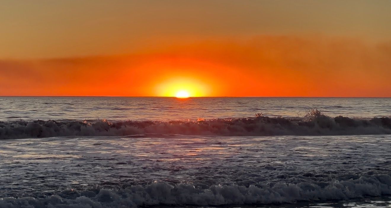 Vibrant ocean sunset with fiery orange sky, glowing sun on horizon, and crashing waves on sandy beach.