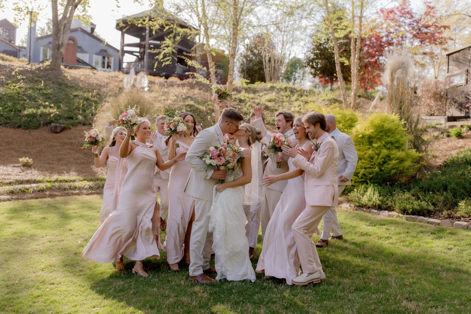 A wedding party in light pink dresses stands together laughing on a sunny spring day in a garden setting.