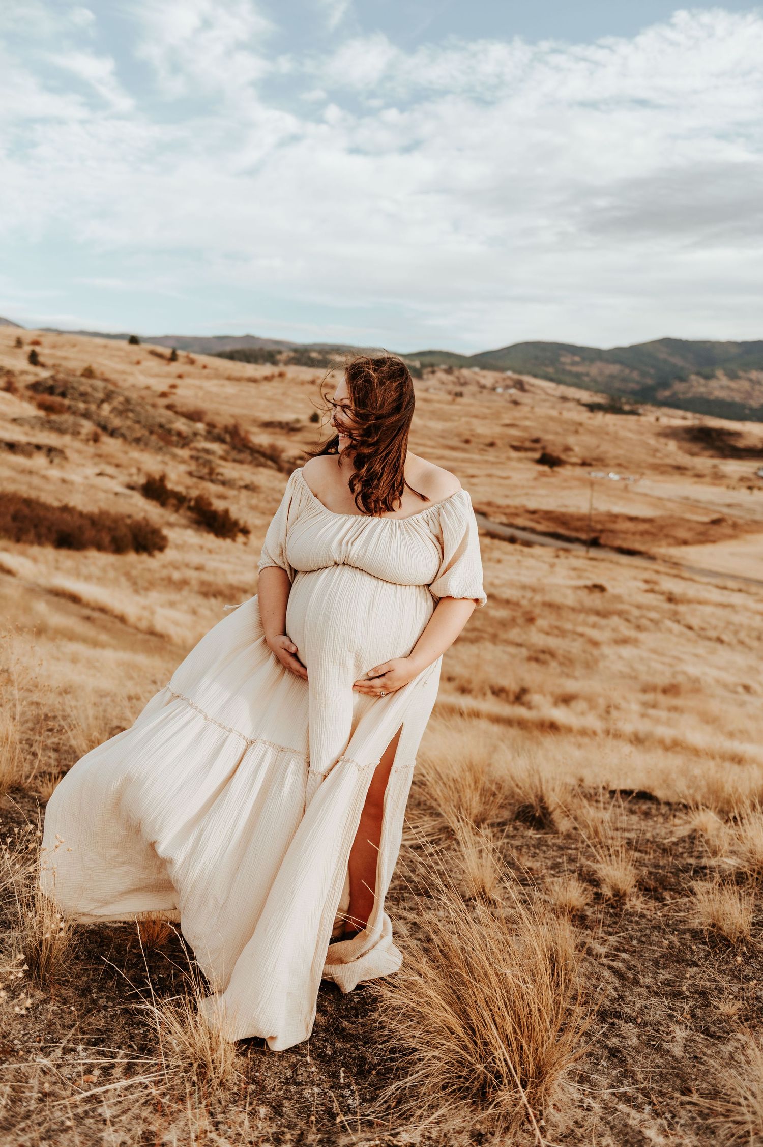 A pregnant woman in flowing white dress poses against a scenic desert landscape with rolling hills.