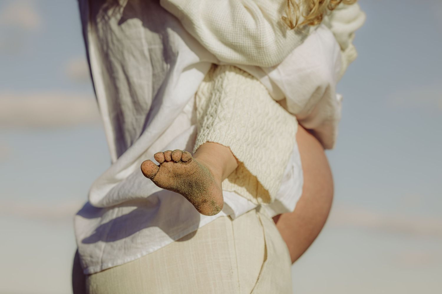 Two people embrace in white clothing at the beach with the ocean and cloudy sky in the background.