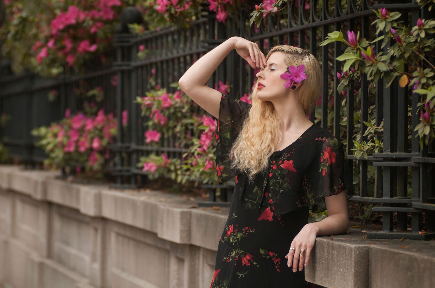 Blonde woman in black floral dress posing near iron fence with pink flowers.