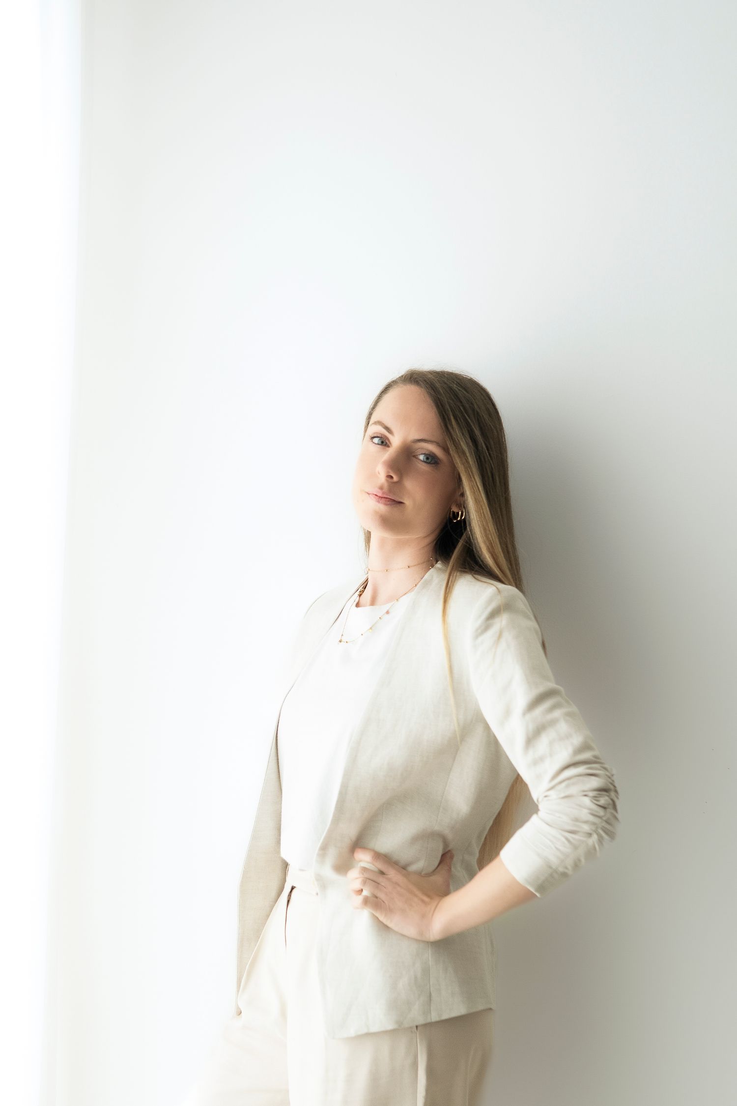 Giovane donna con abiti professionali beige, appoggiata a una parete bianca luminosa, guarda dritta l'obiettivo tenendo una mano sul fianco