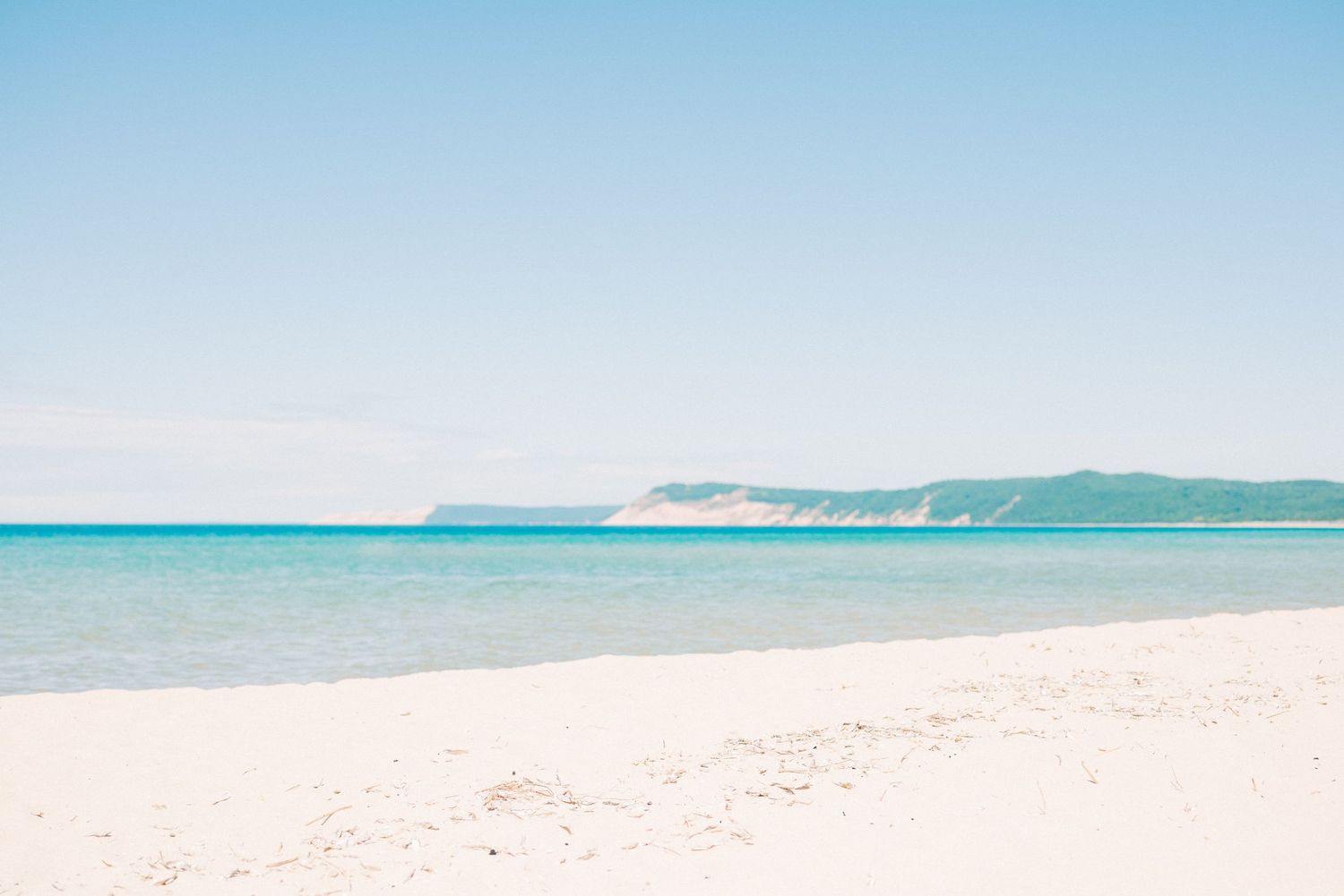 Tranquil beach scene with crystal clear turquoise waters and distant green hills under soft blue skies.