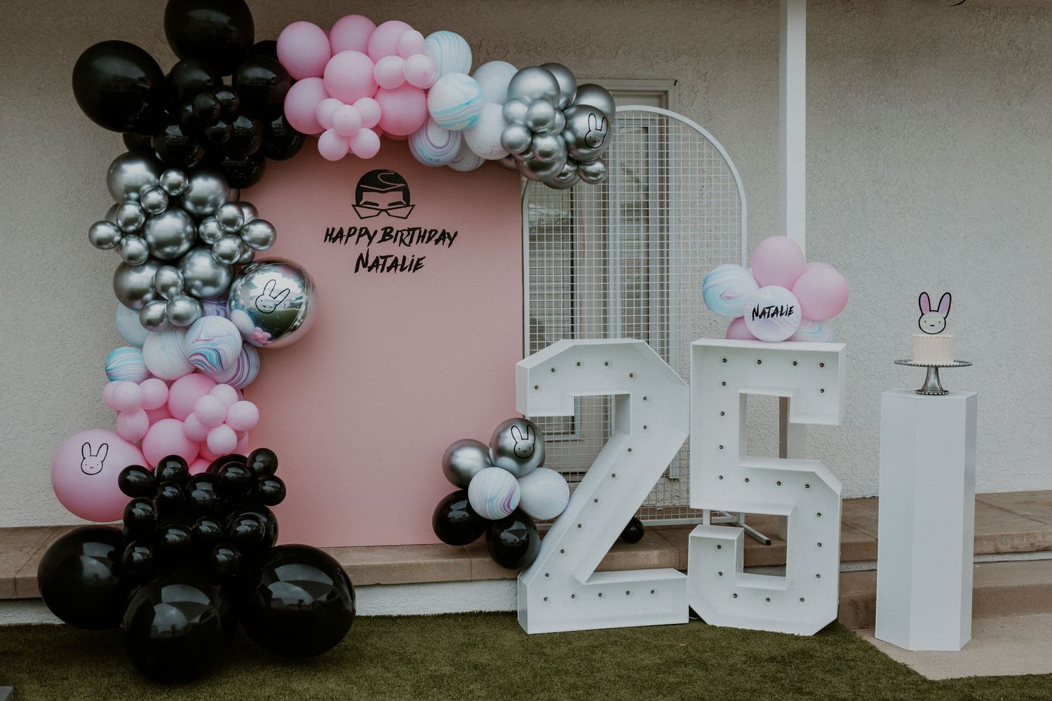 A pink backdrop with black, silver and pink balloon arch and large white number 25 display for a celebration.