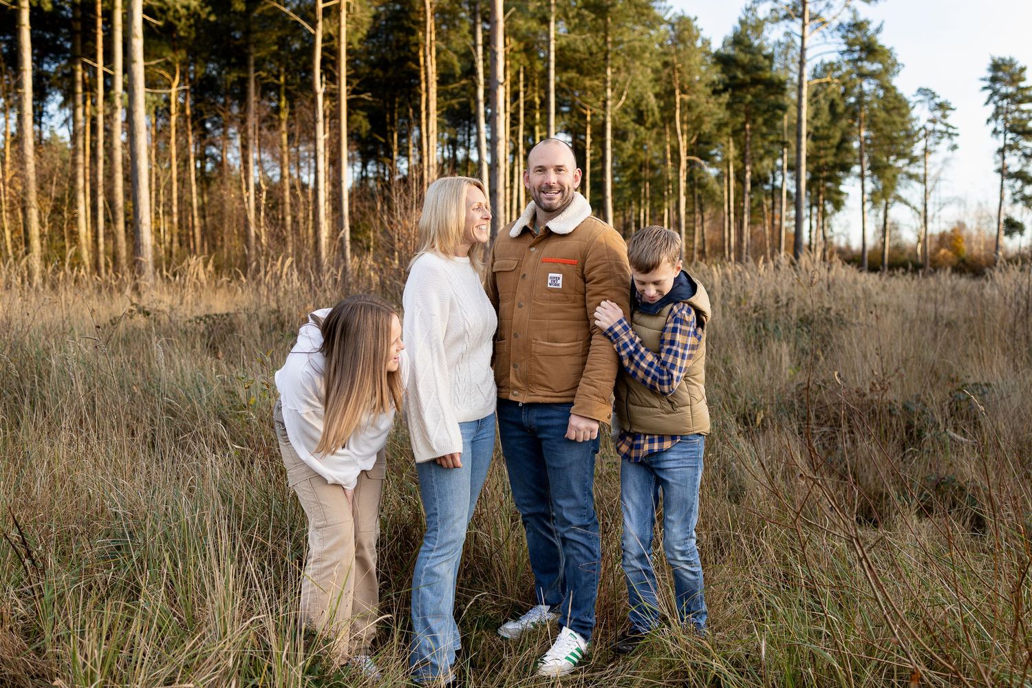 Family of four poses together in casual attire among tall pine trees at sunset.