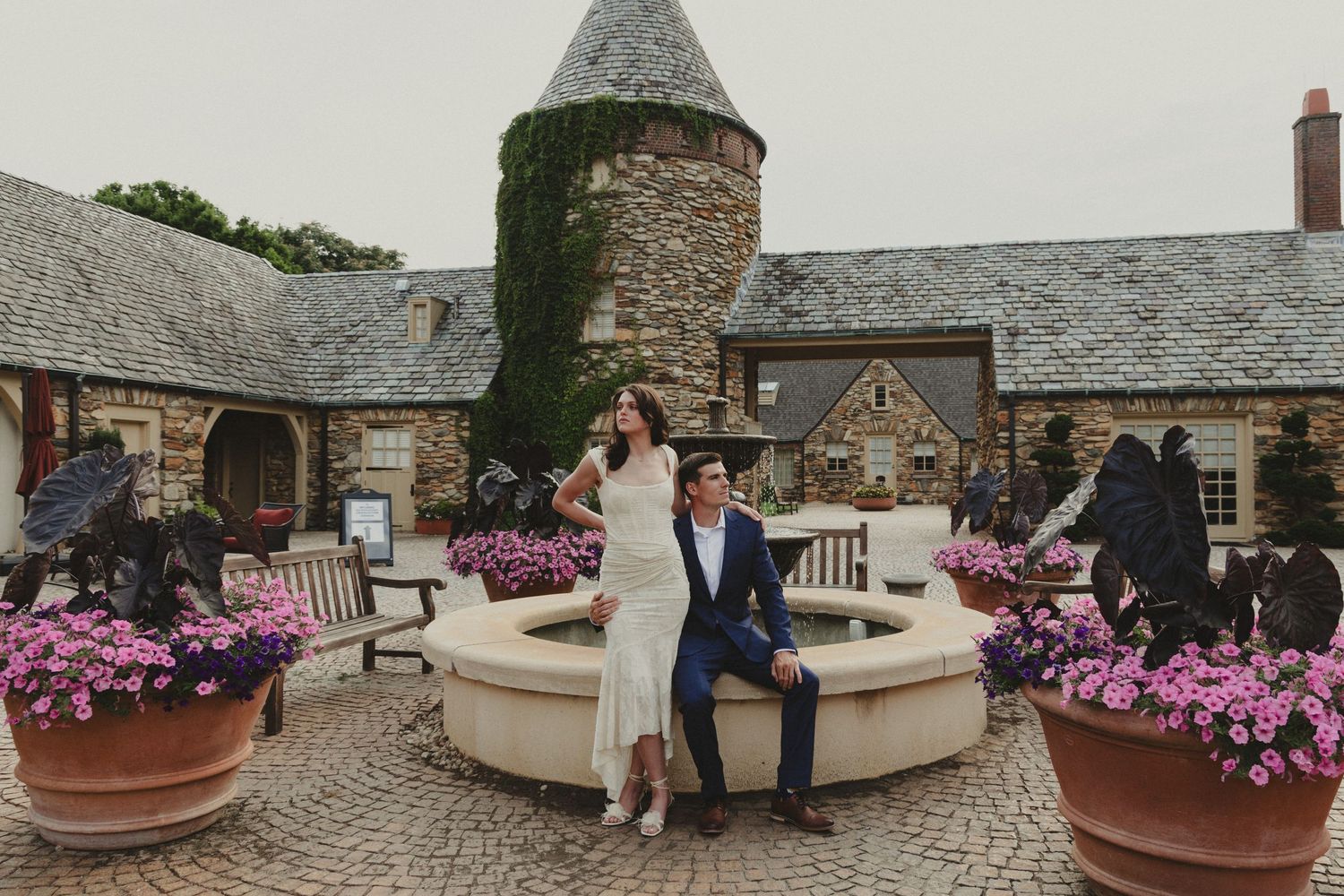 Wedding couple seated on curved stone bench surrounded by pink flowers at rustic stone venue.
