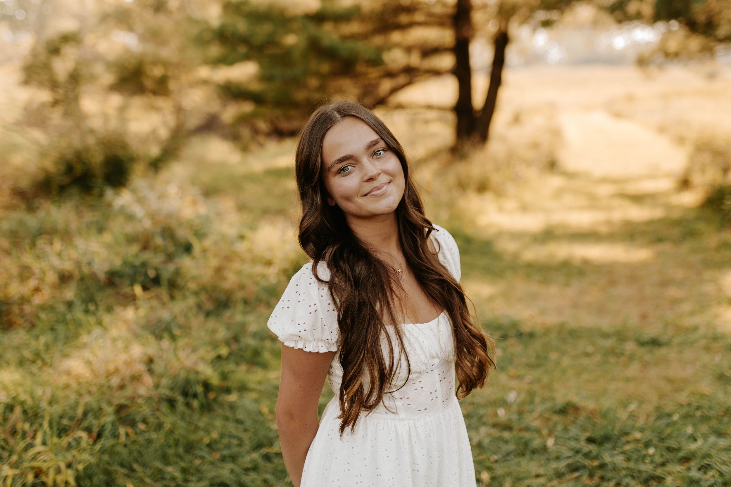 A person in a white lace dress stands in a sunny field with trees in the background during golden hour.