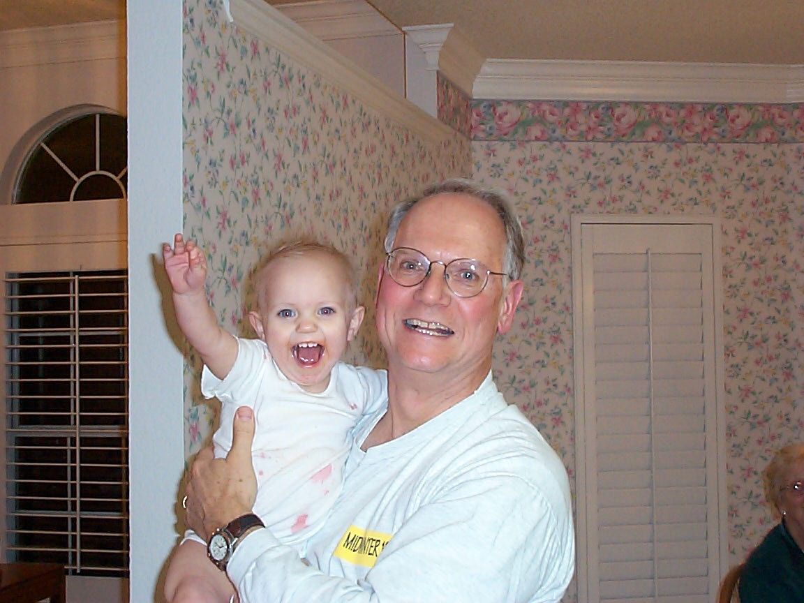 A smiling pair in a room with floral wallpaper and white shuttered windows visible in the background.