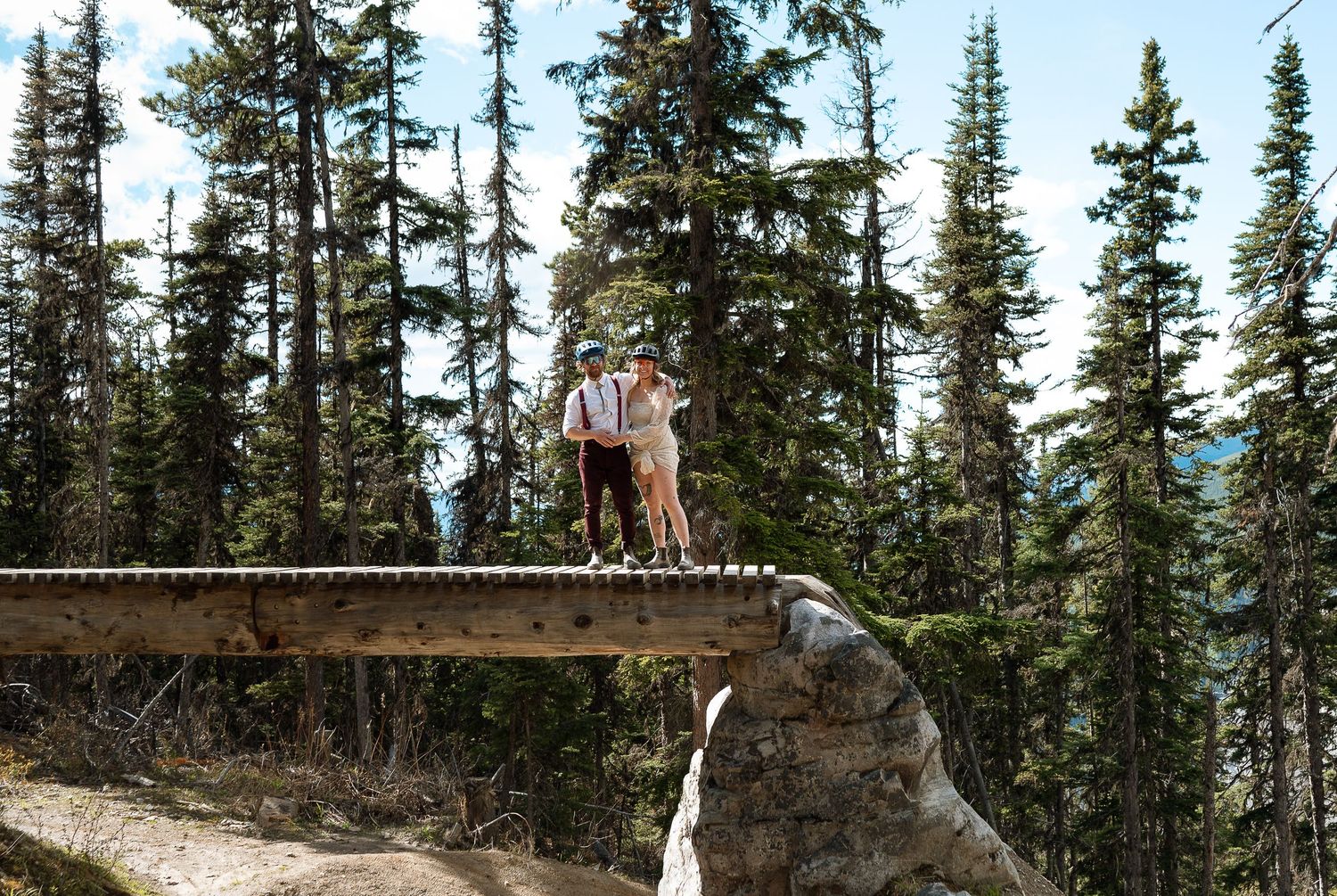 Mountain bikers stand on a wooden platform overlooking a scenic forest landscape with evergreen trees.