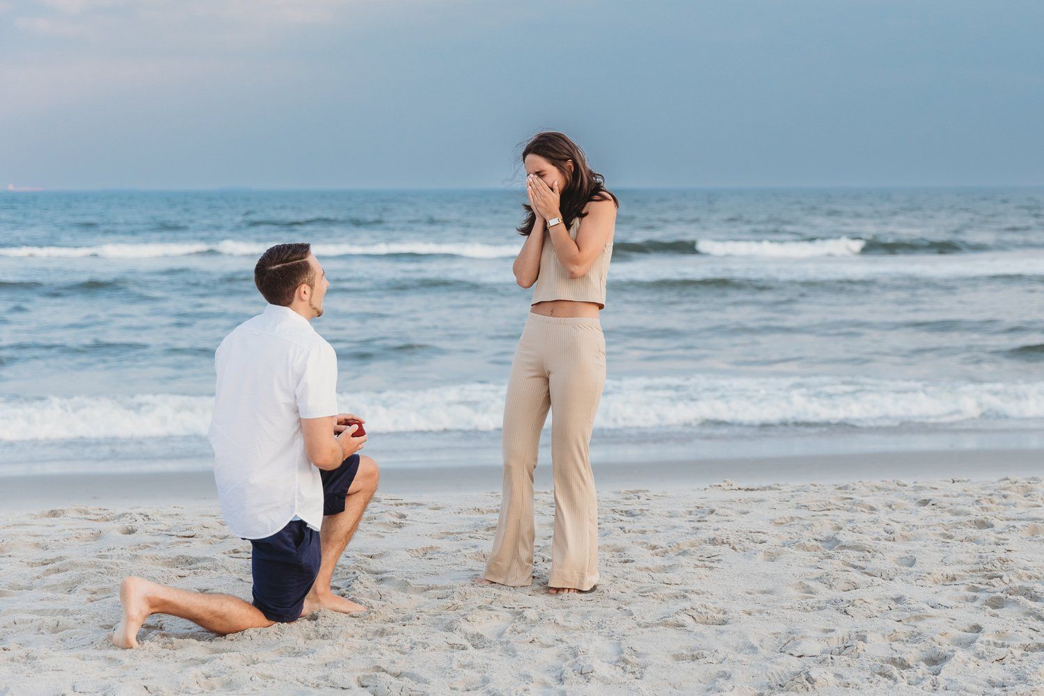 A romantic beach proposal scene with waves crashing in the background during sunset.