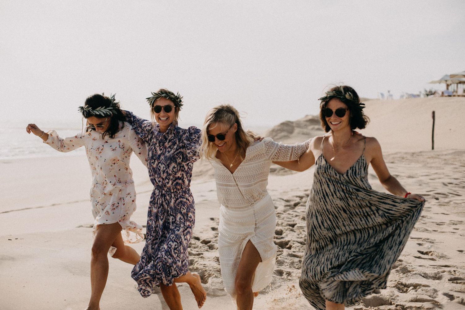 Four friends dancing and twirling on a sandy beach in flowy dresses during a carefree summer moment.