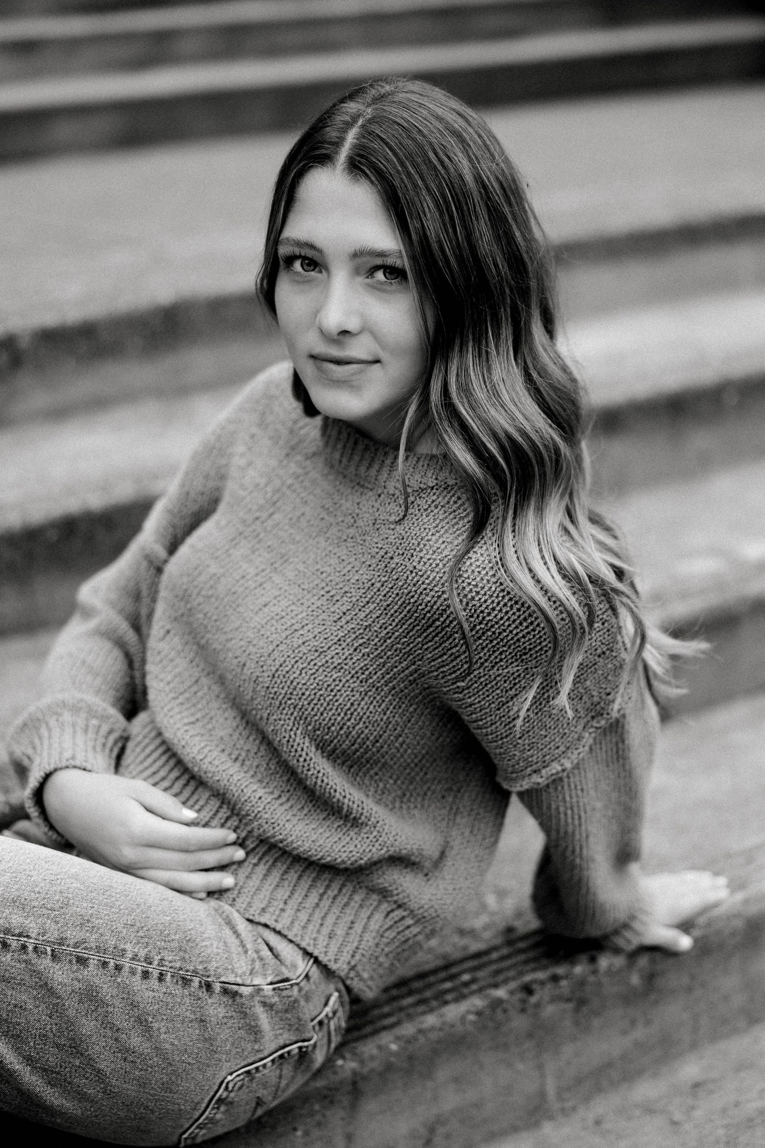 Black and white portrait of a teenage girl sitting on steps in a sweater and jeans.