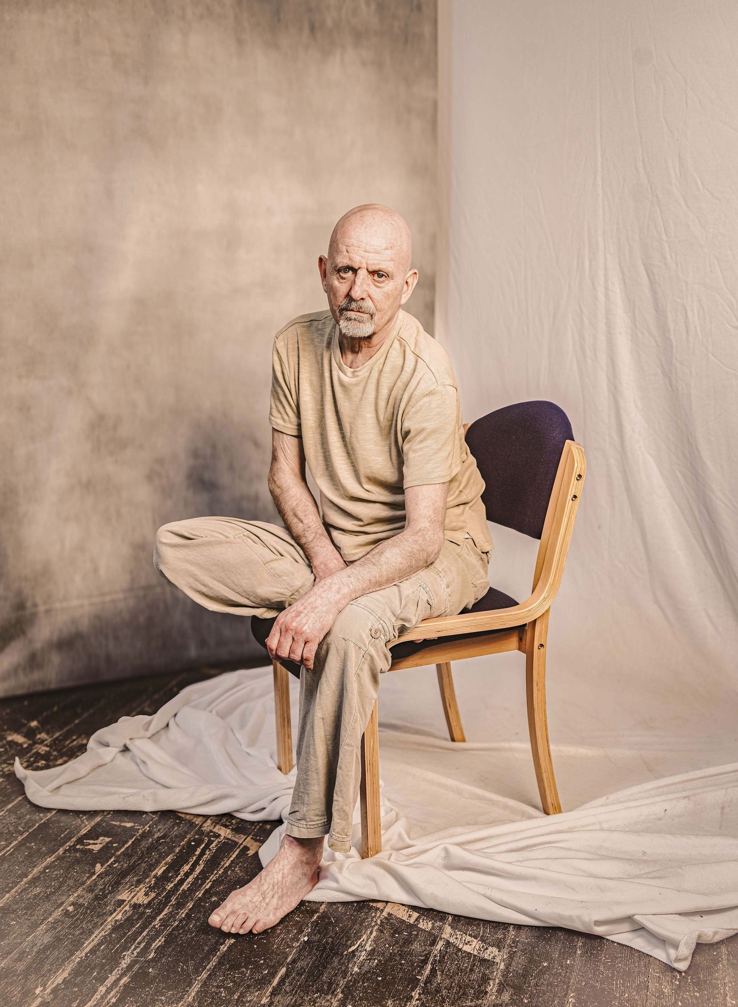 Figure in beige clothing sits cross-legged on wooden chair against neutral backdrop with white fabric draped on floor.