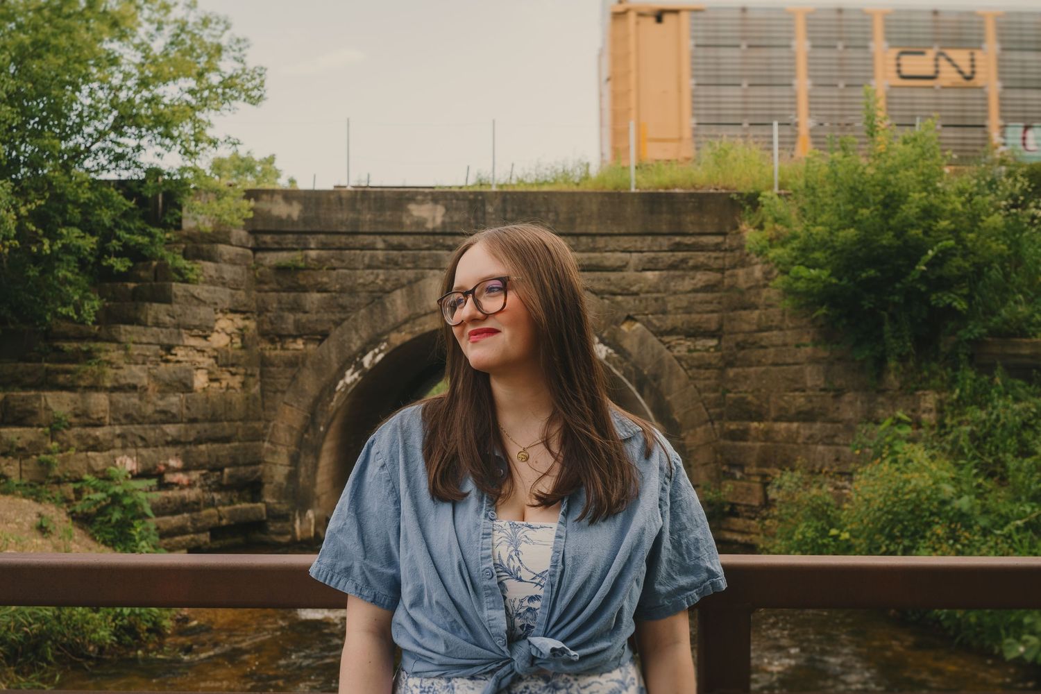 Someone wearing glasses and tied denim top sits on wooden railing near stone wall on sunny day, looking to side and smiling.