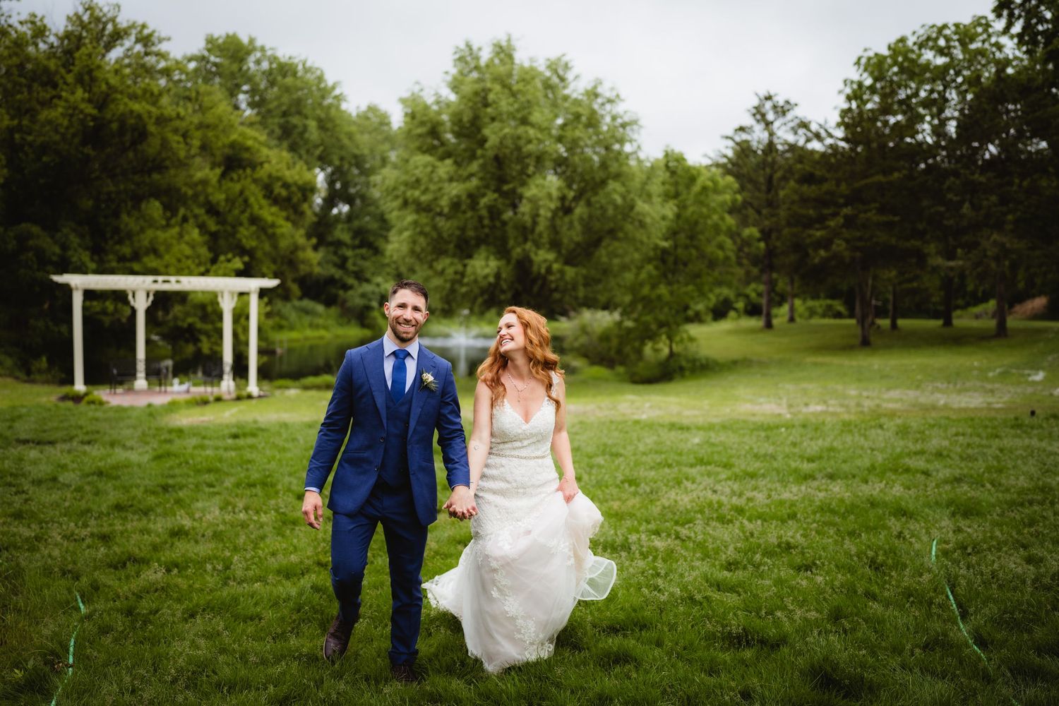 Couple walking hand in hand across green lawn with white gazebo in background.