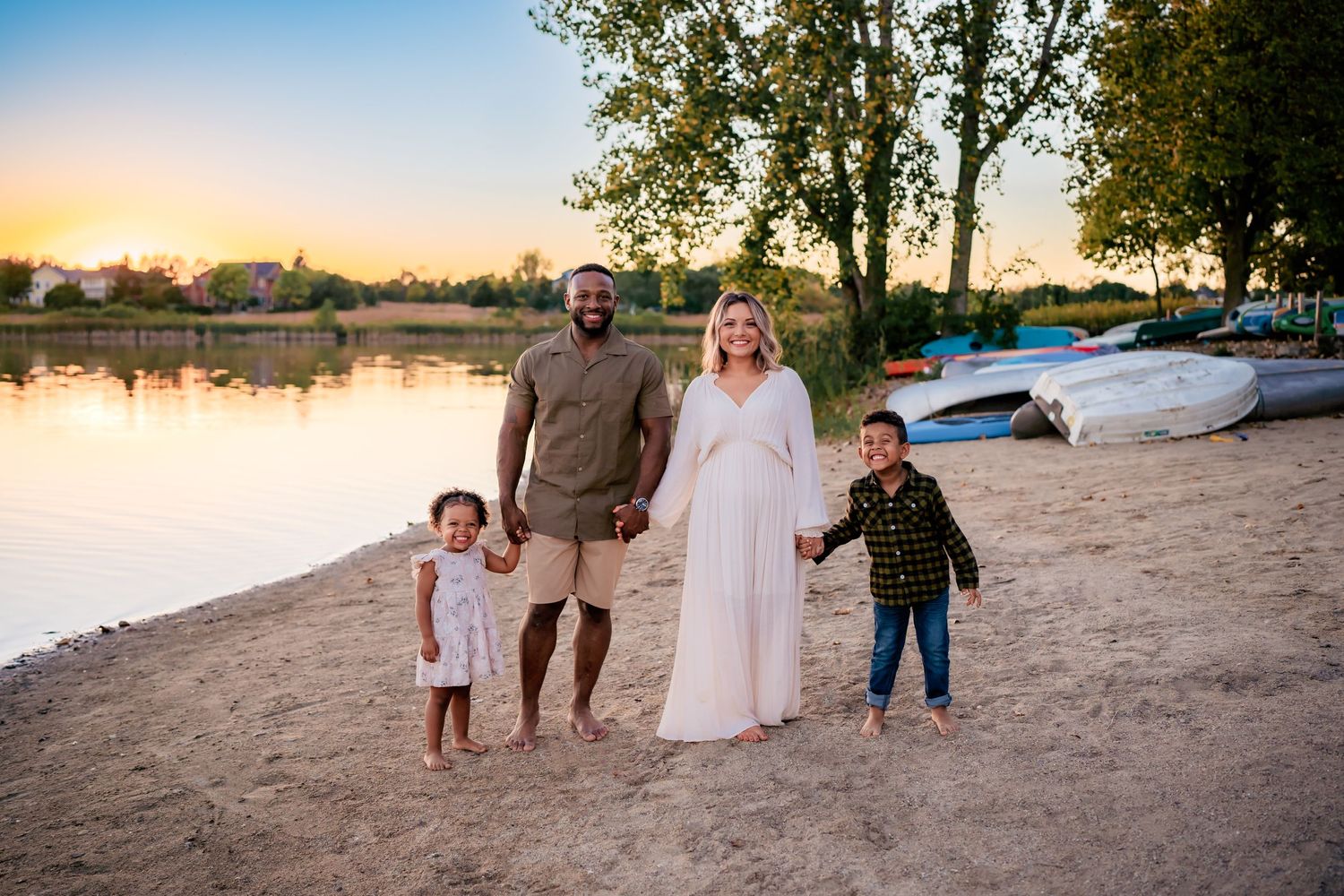 Family of four poses on a sandy beach at sunset with a serene lake and boats in the background.