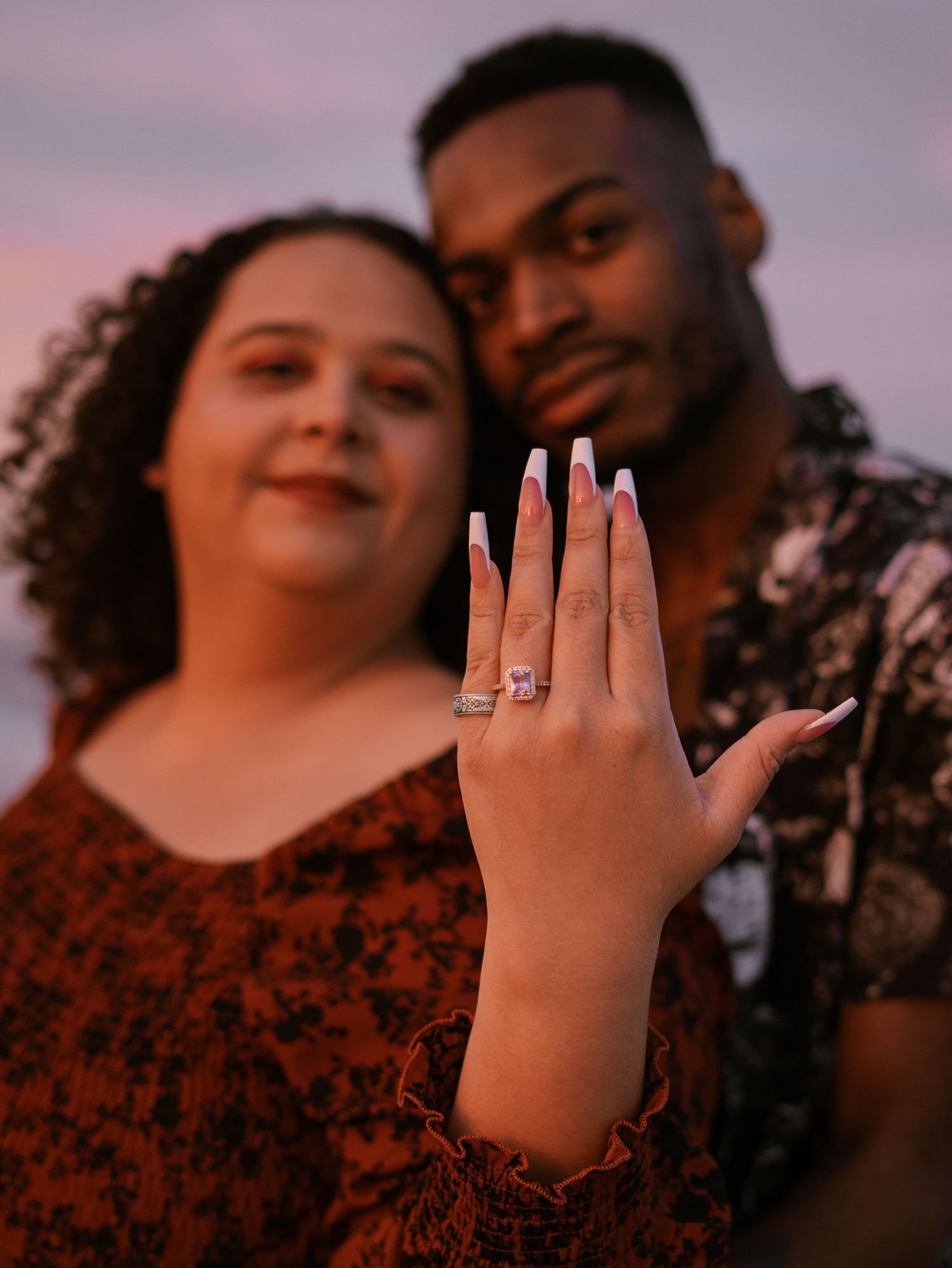 A hand showing off an engagement ring at sunset while a couple in red and floral prints pose in the background.