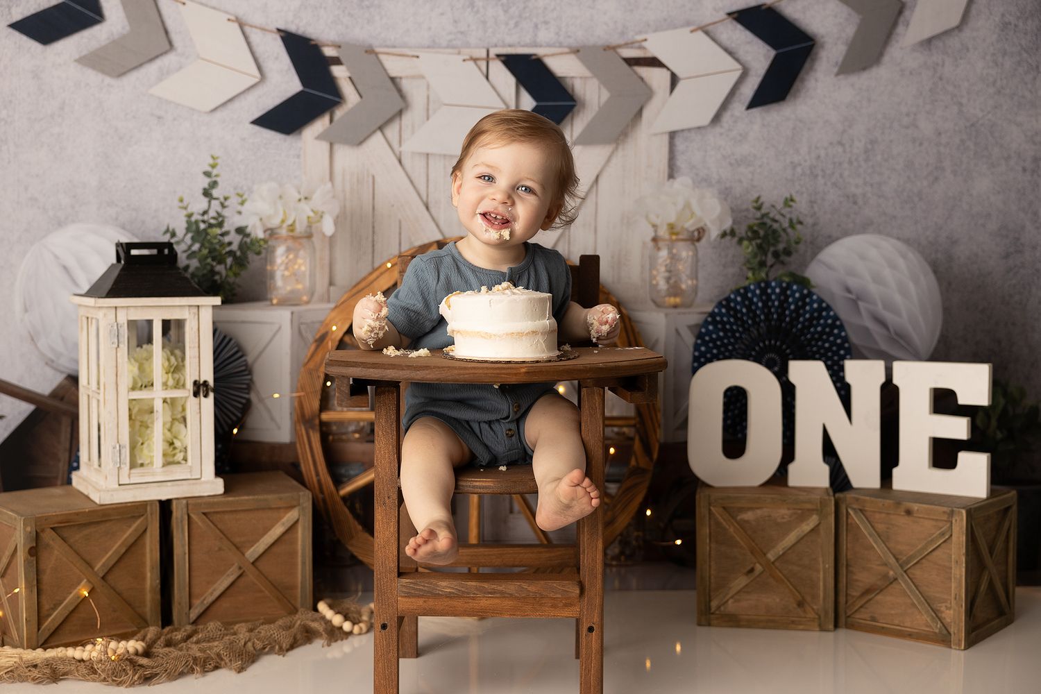 Vintage styled first birthday cake smash setup with wooden boxes, lantern and decorative 'ONE' letters against chevron backdrop.