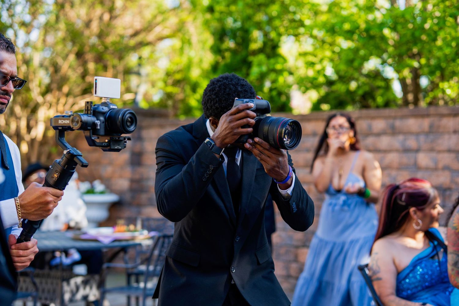 Professional wedding photographers in black suits working with cameras at an outdoor event with guests in formal attire.