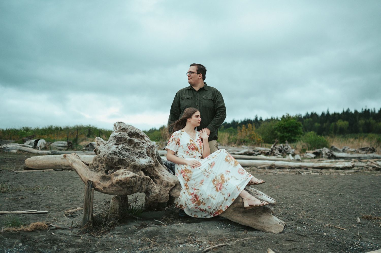 Person in floral dress poses on driftwood logs at misty Pacific Northwest beach under moody cloudy skies at sunset.