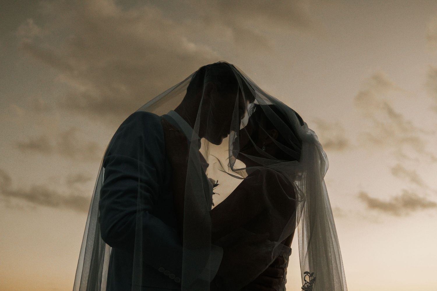 Silhouetted couple sharing a romantic kiss under flowing wedding veil at sunset with dramatic sky in background.