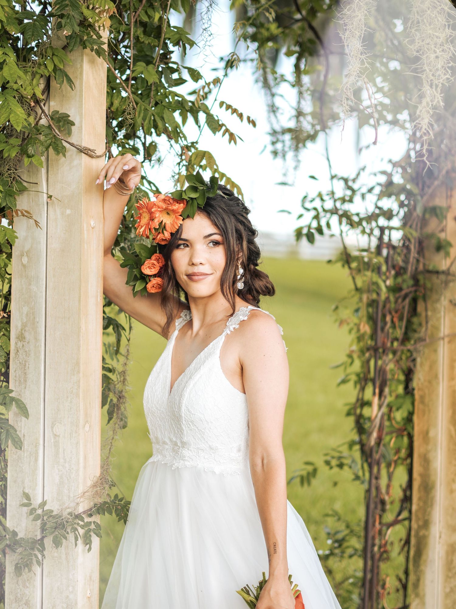 A bride in a white lace dress holds a coral and peach bouquet amid green foliage in an outdoor garden setting.