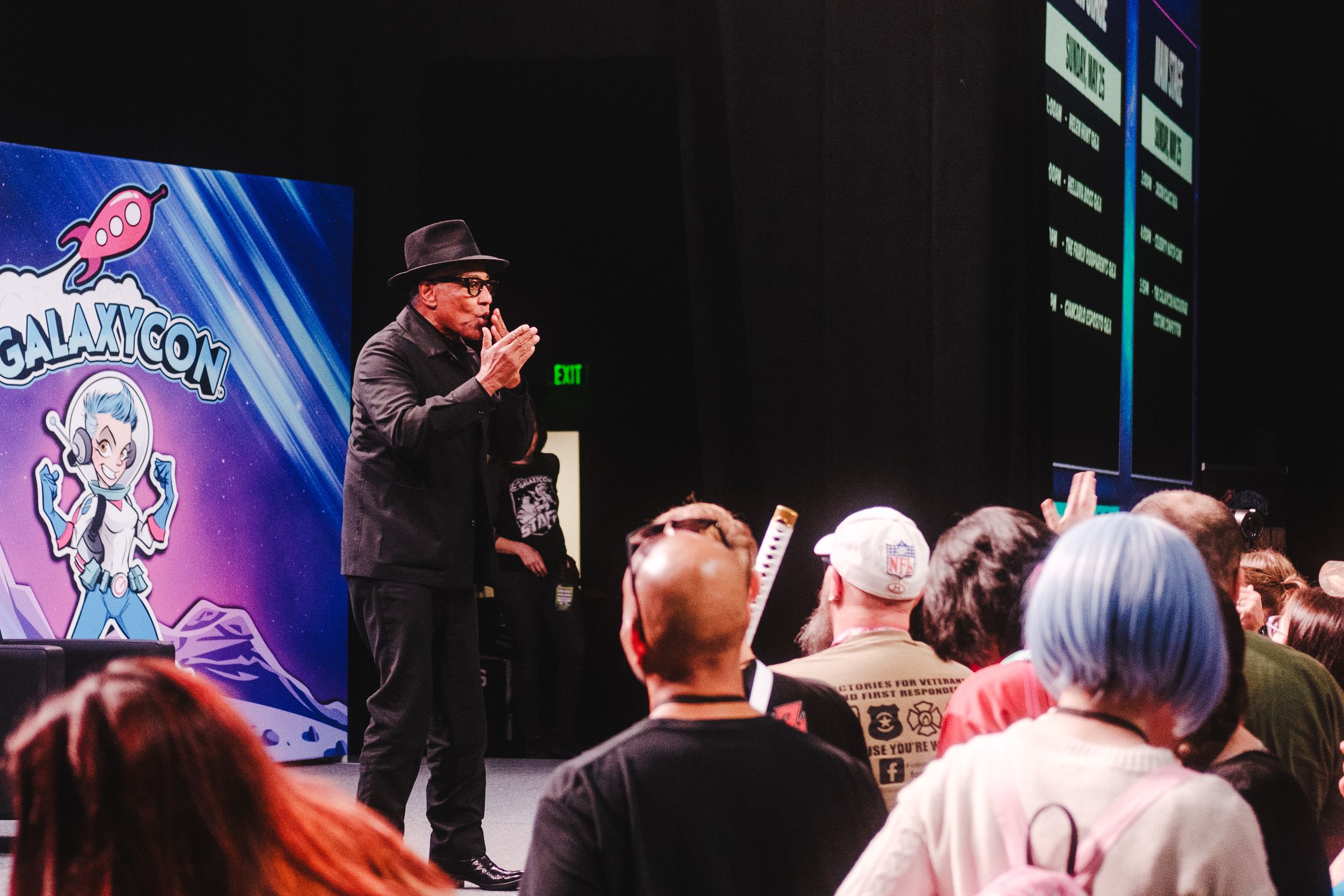 A performer speaks into a microphone on stage while audience members watch at a dark indoor event venue.