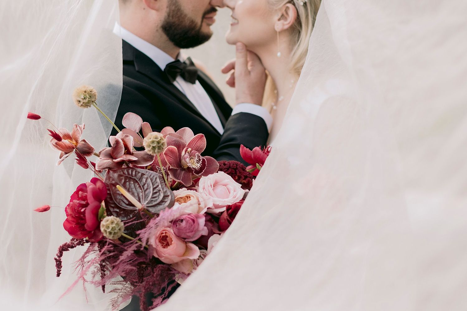 Couple shares an intimate moment behind a stunning deep red and pink bridal bouquet under a veil.