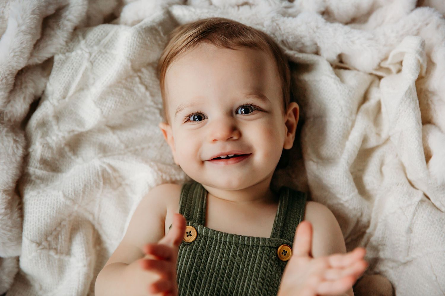 A happy baby in green overalls lying on soft cream-colored blankets.