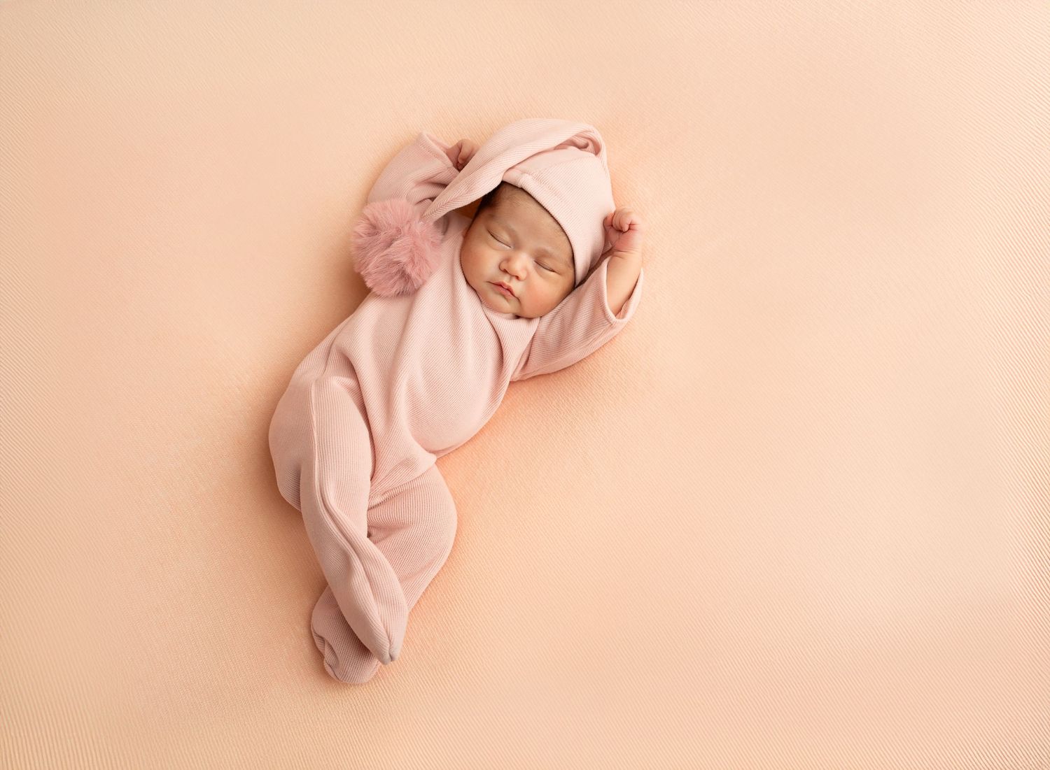 Peaceful sleeping newborn baby wearing soft pink onesie and hat resting on peach-colored backdrop.