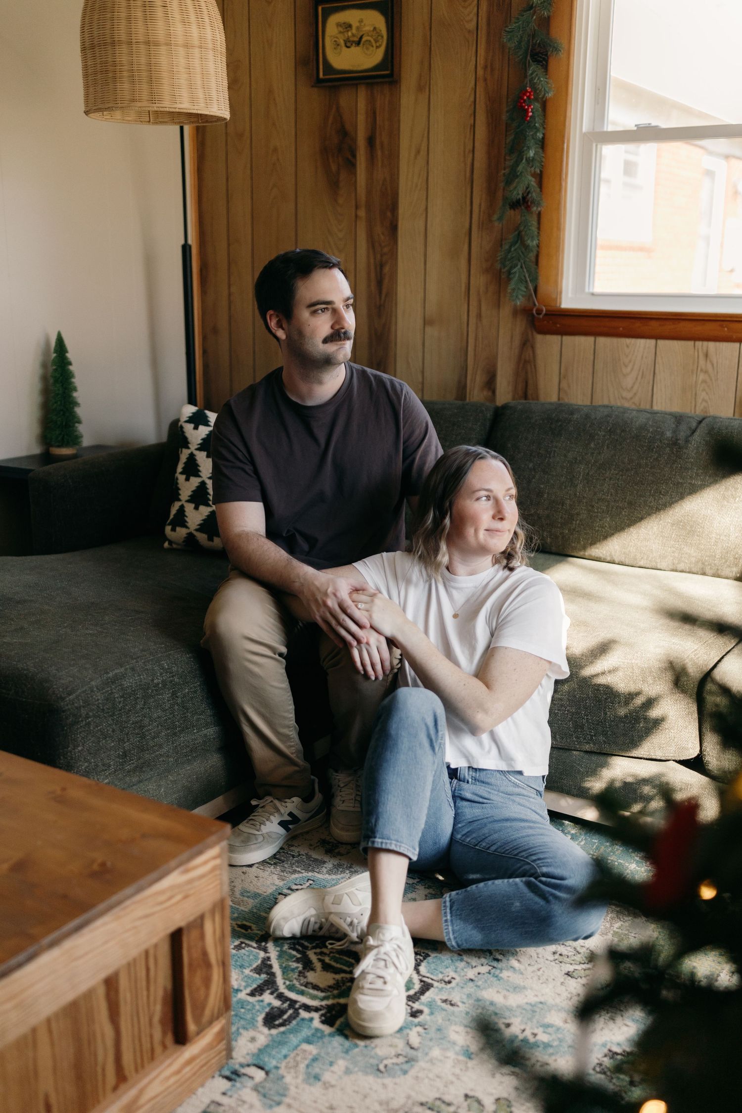 A couple relaxes on a gray couch in a rustic room with wood paneled walls and pendant lighting.