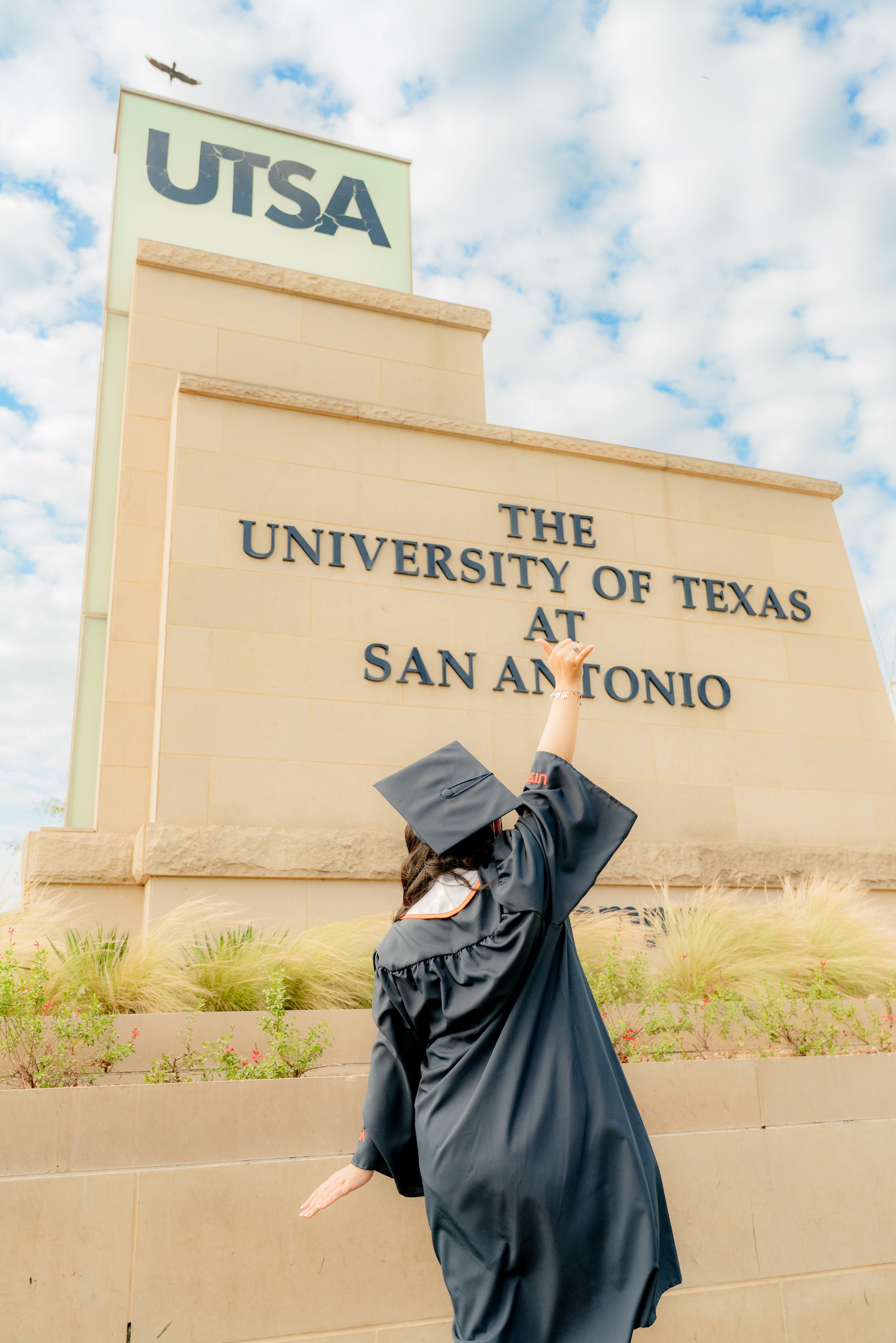 Graduation photo at UTSA campus sign with cap and gown silhouette.