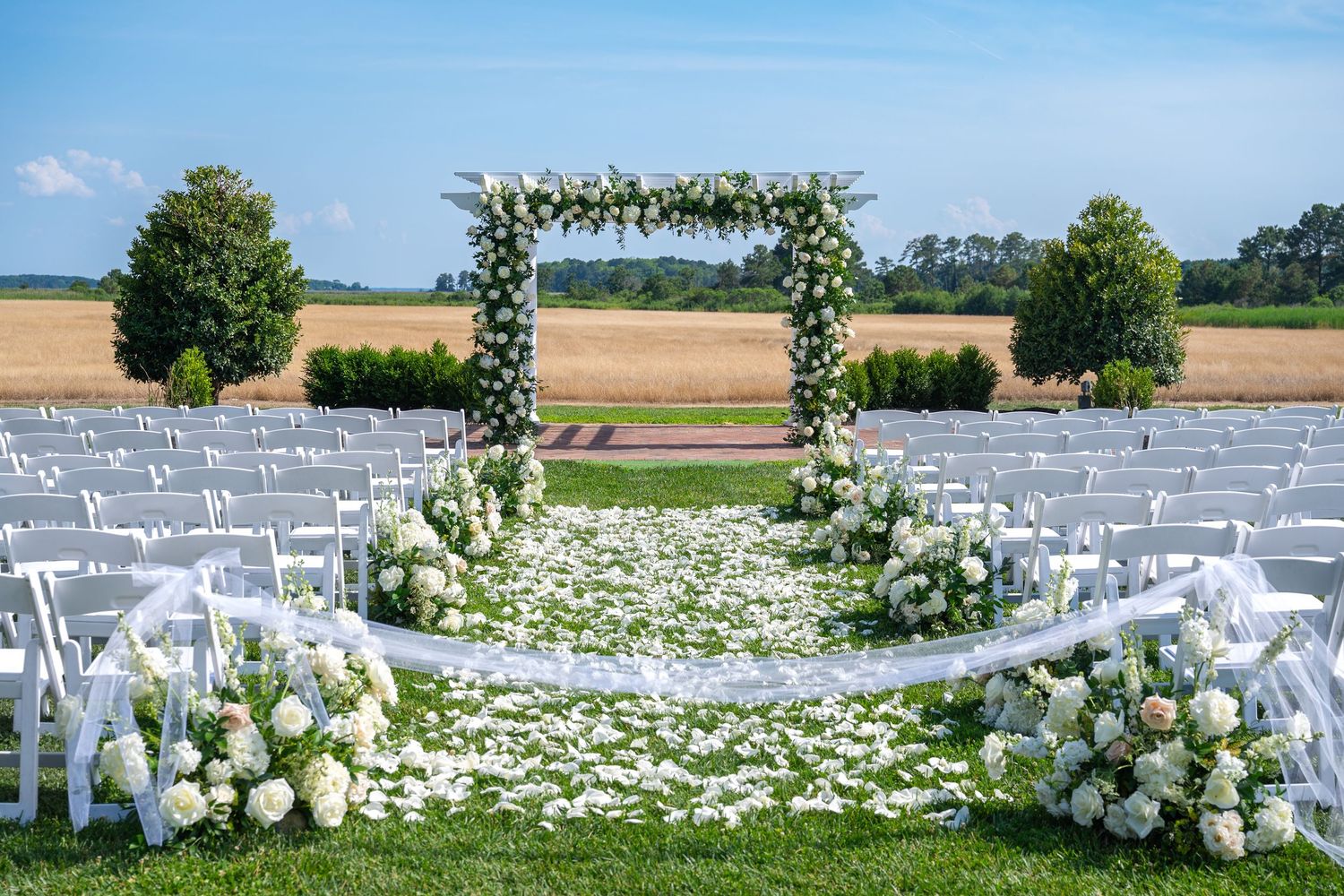 Outdoor wedding ceremony setup with floral arch, white chairs and rose petals on lawn overlooking a golden field.