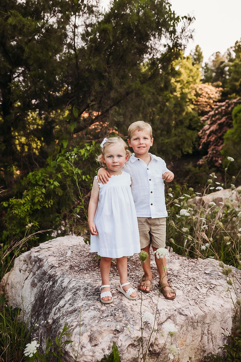 Two small children stand together on a large rock formation in an outdoor garden setting with blooming flowers.