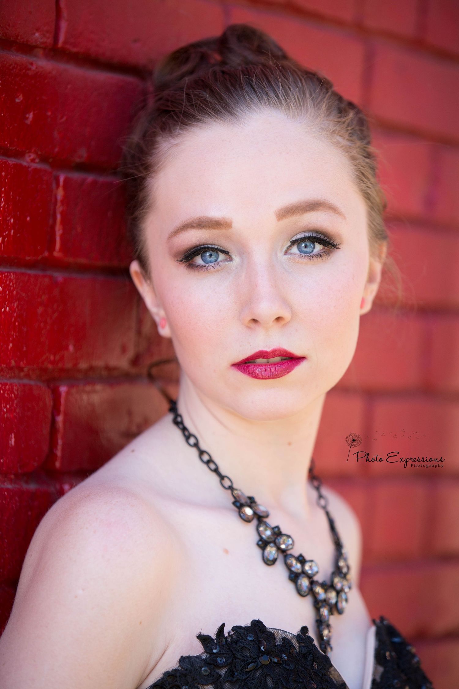 Portrait against red brick wall featuring dramatic makeup and black statement necklace.
