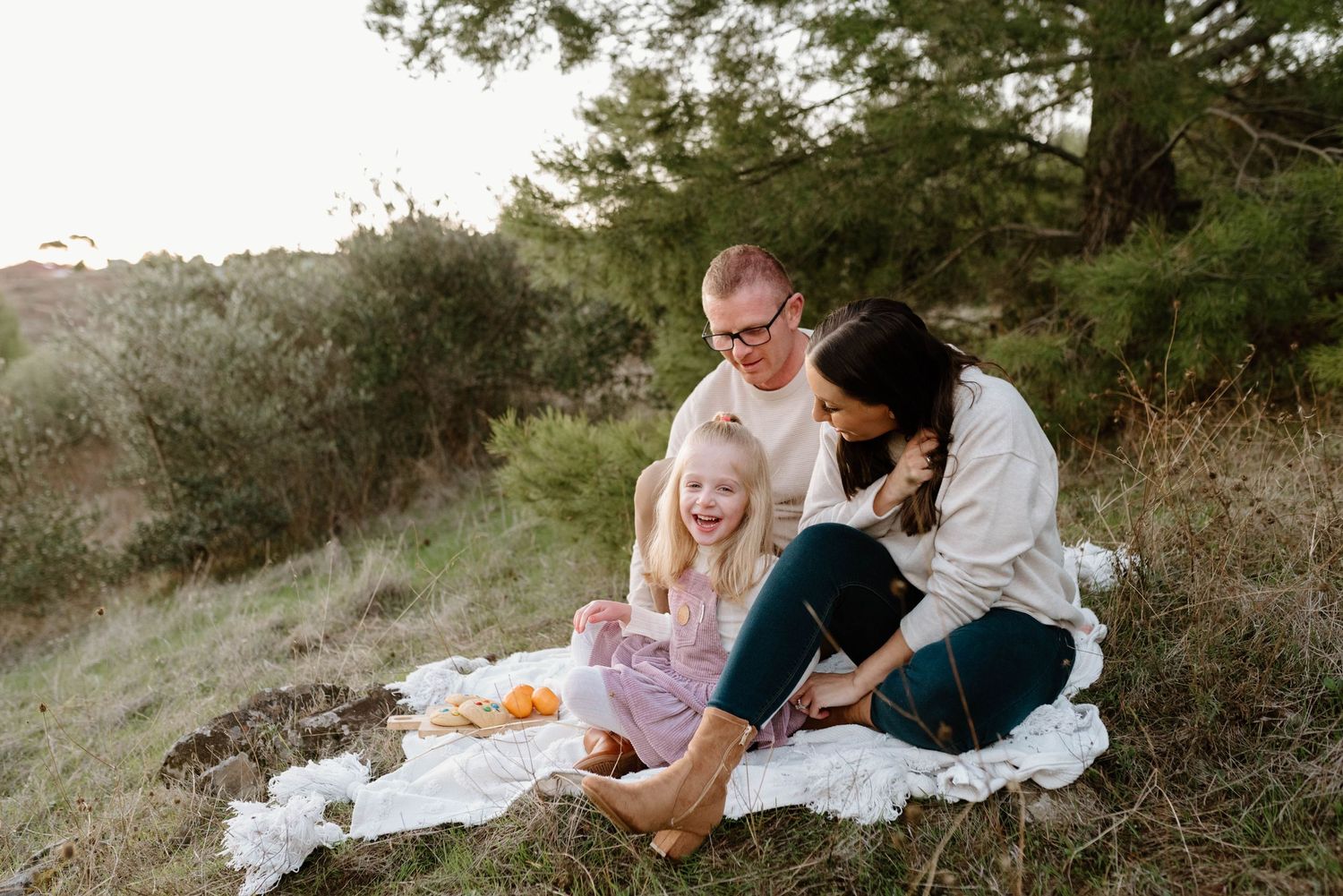 Family having picnic on white blanket during sunset in grassy hillside setting.
