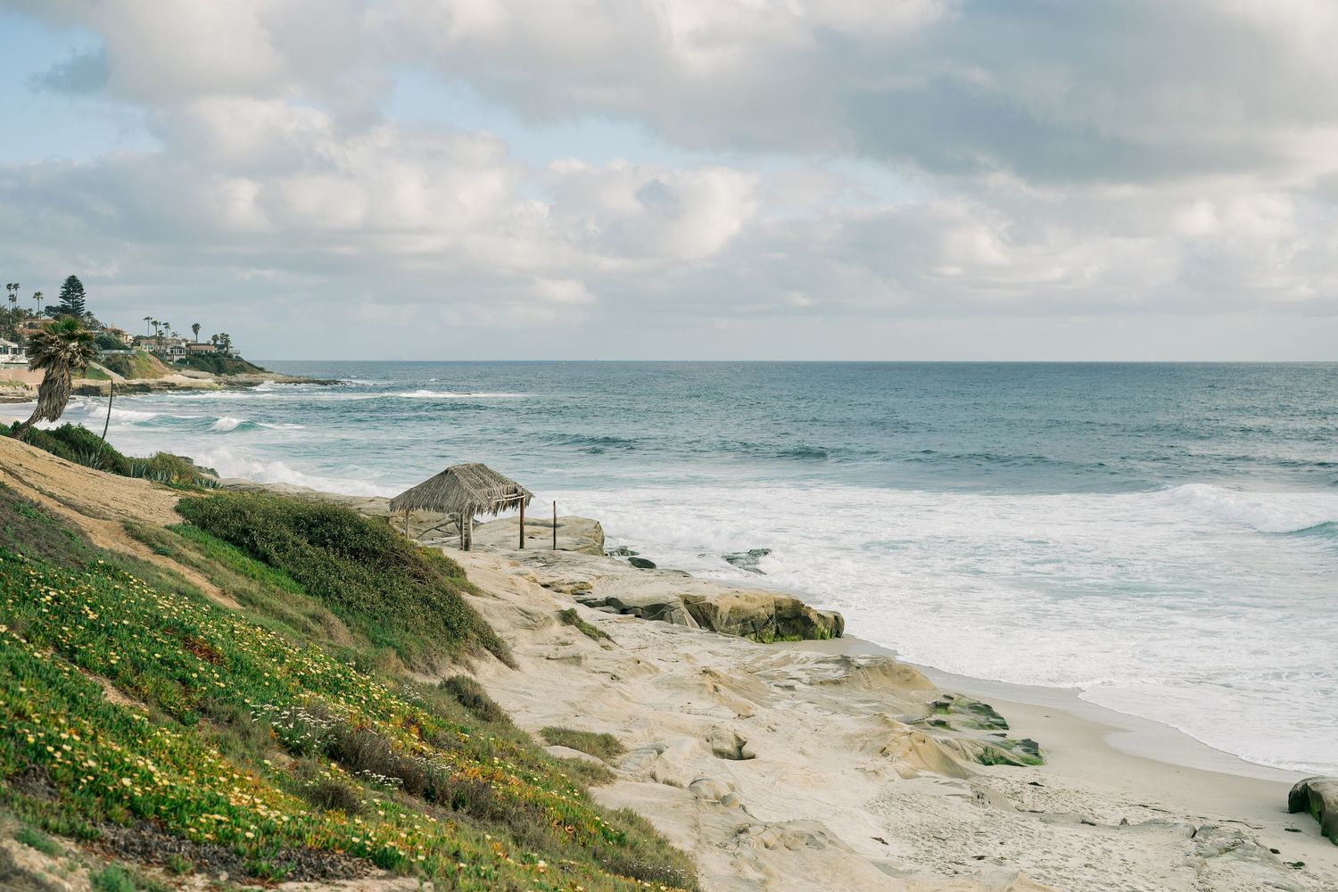 Coastal landscape with thatched palapa on sandy beach overlooking rough ocean waves and green hillside in Mexico.