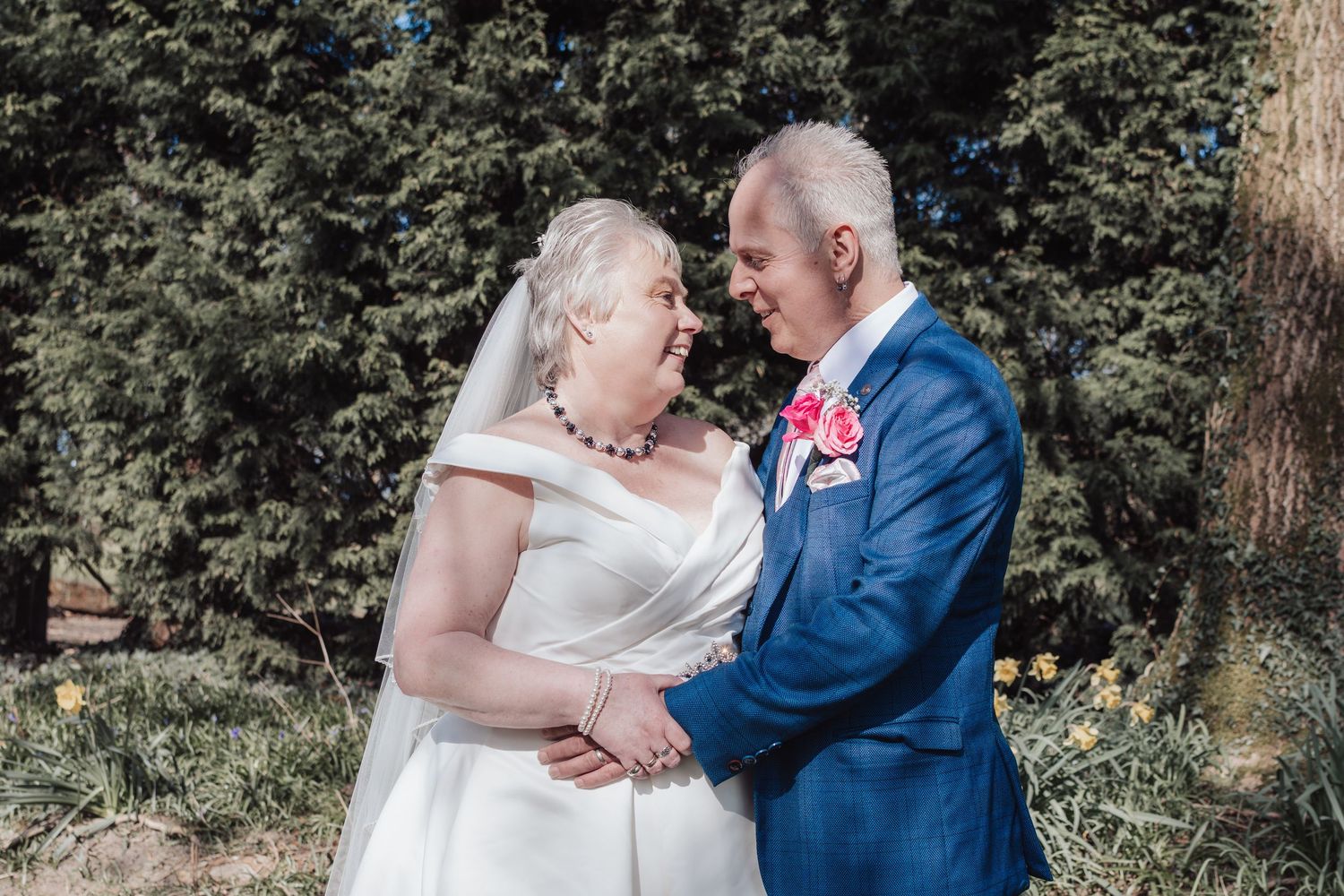 A couple shares a tender moment in an outdoor garden setting wearing elegant wedding attire.